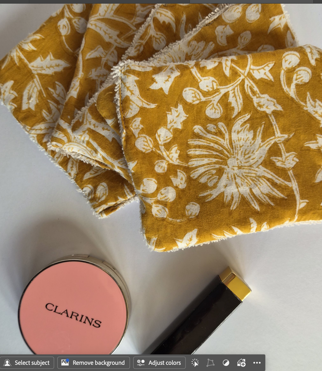 Cosmetic wipesmustard yellow fabric items with a white floral block-print pattern, displayed on a white surface alongside a pink Clarins compact and a black and gold cosmetic tube.